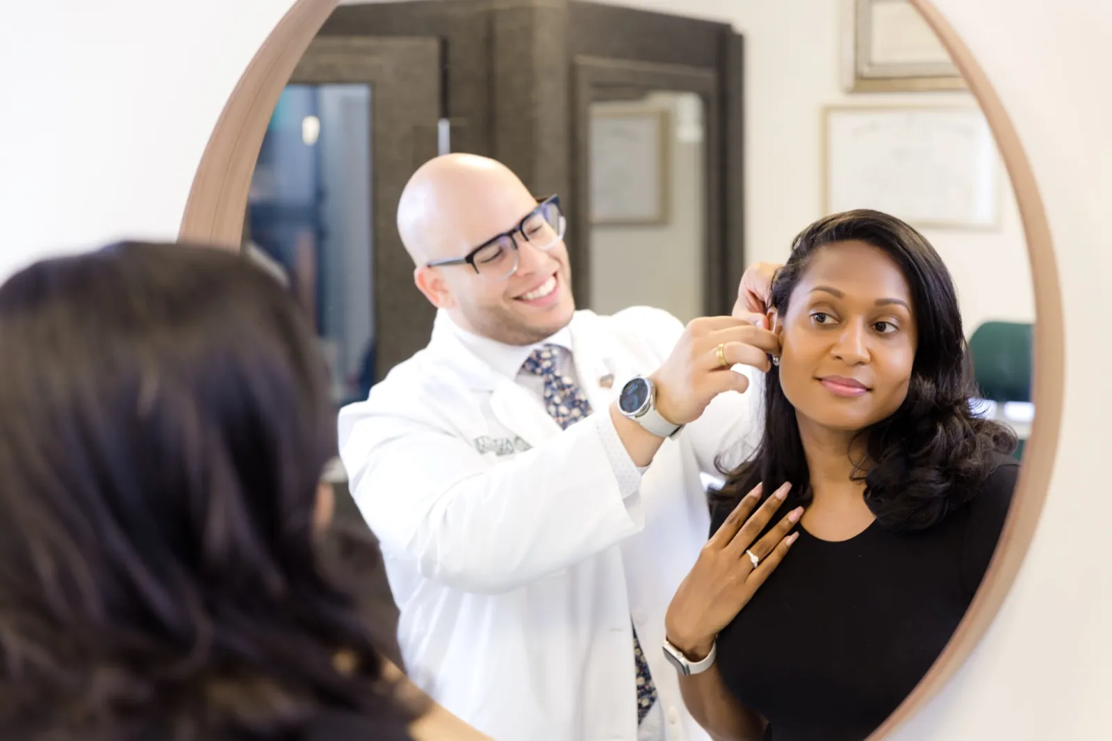 Jaysee fitting a hearing aid on a smiling patient in front of the mirror