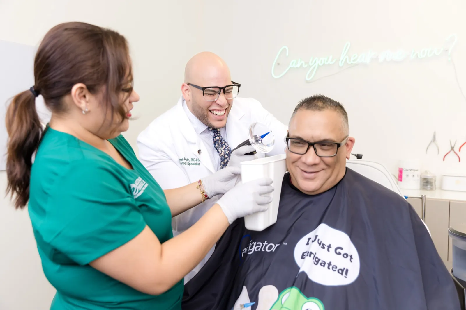 A patient smiling under the Earigator cape as Jaysee and a Patient Care Coordinator clean his ears