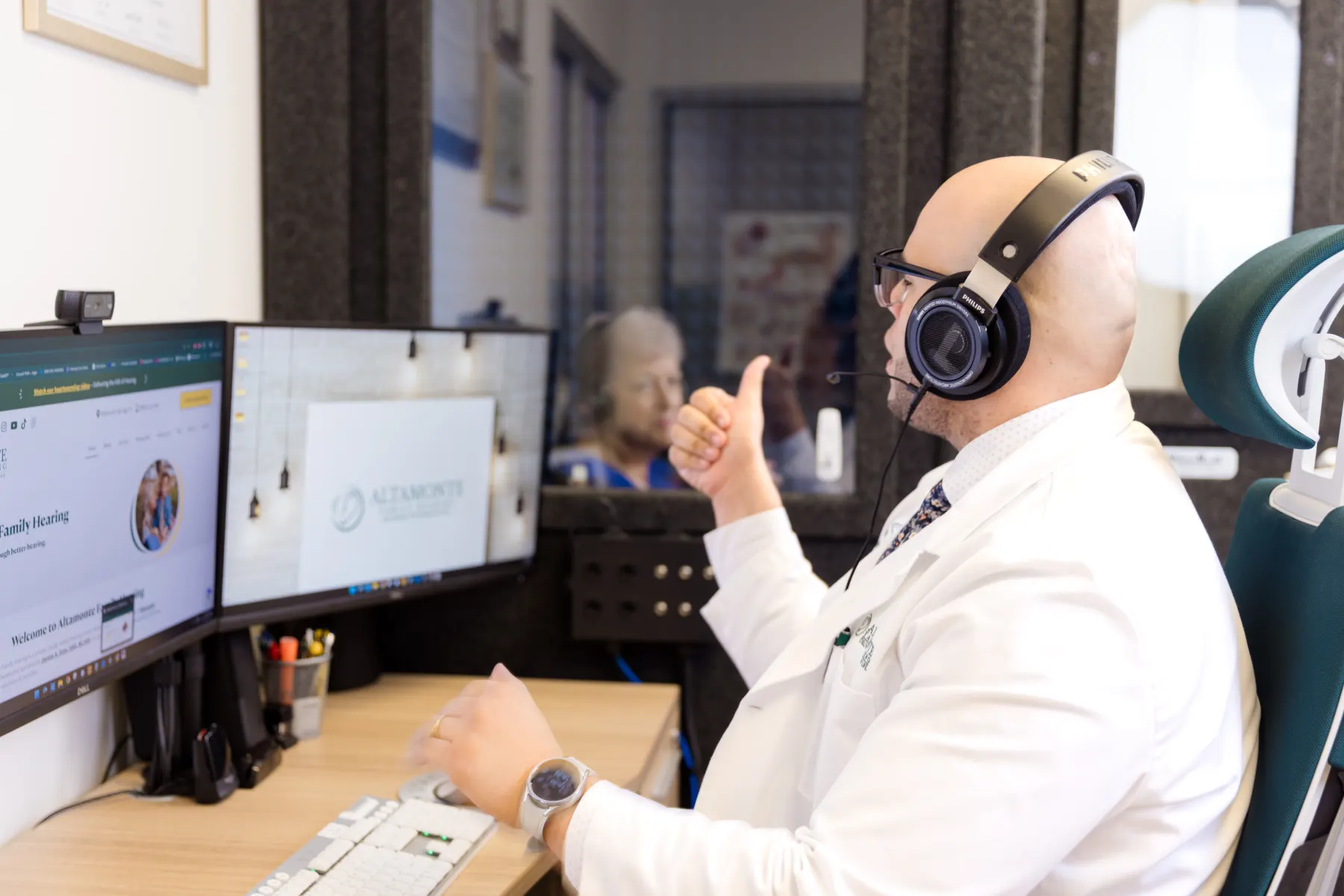 Jaysee giving a thumbs-up at his desk in headphones while the patient sits in the sound booth