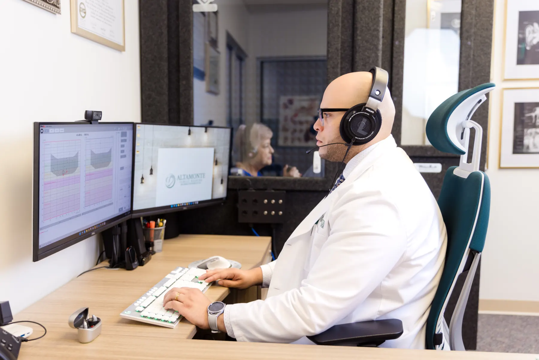 Jaysee running a hearing test from his desk while the patient sits in the sound booth