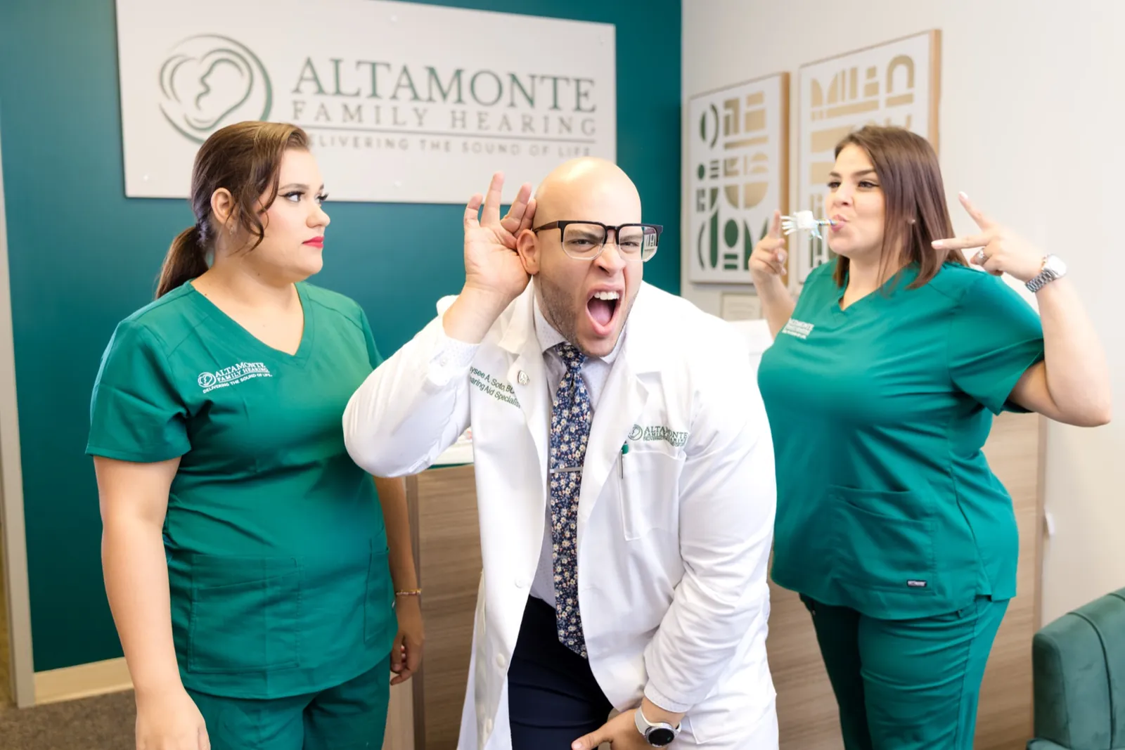 Jaysee cupping his ear and grinning between Grace and Francesca at the front desk